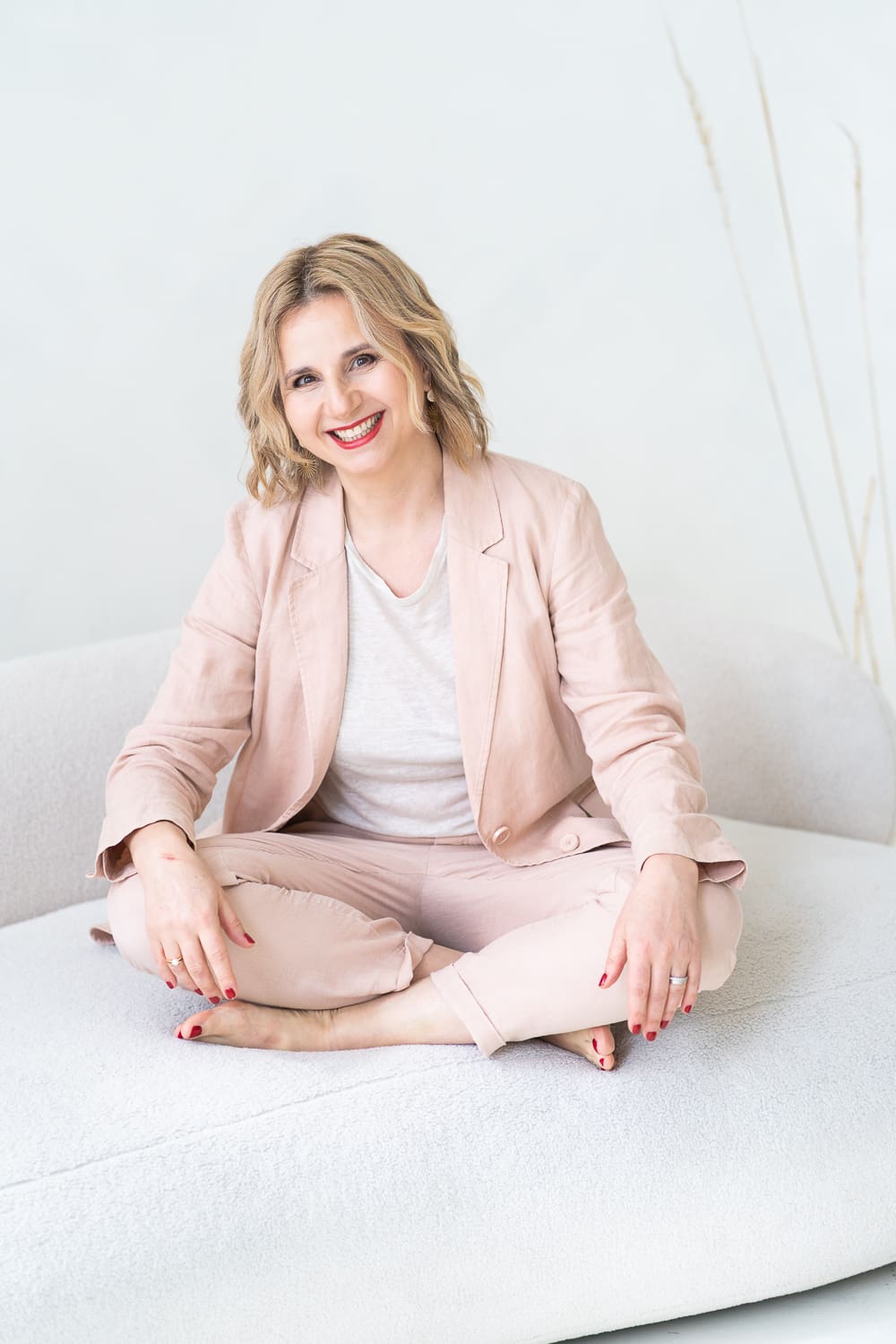 žena v béžovo růžovém kostýmku sedí v tureckém sedu na světlé pohovce a směje se